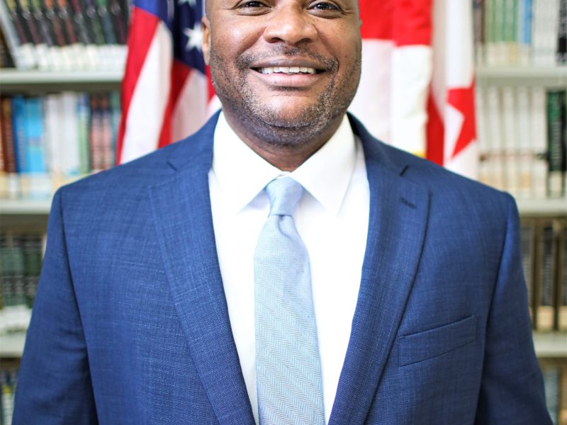 Leonard Bishop, ANC of the D.C. Jail, is posing for a photo in front of two flags, wearing a blue suit and light blue tie.