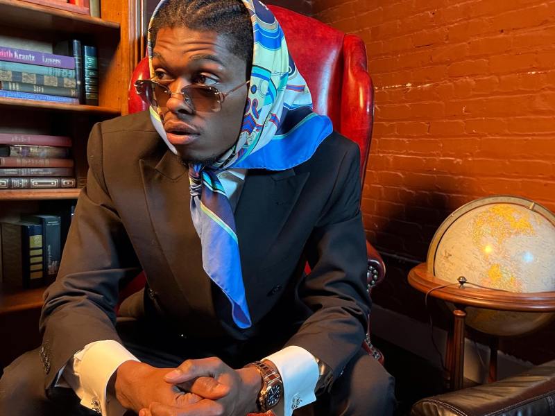 Naeem Khaliq is sitting in a brown leather chair wearing a suit and a colorful scarf on his head