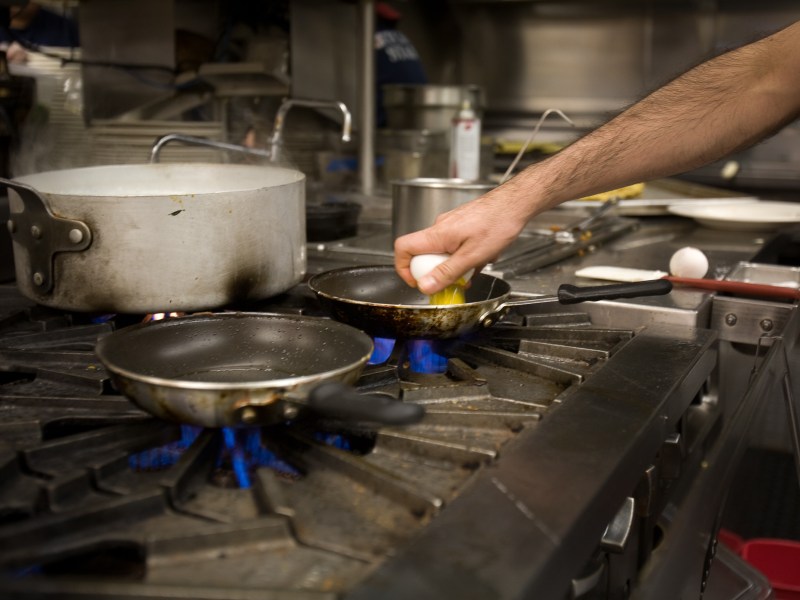 A chef cracks an egg over a pan on a gas stove.