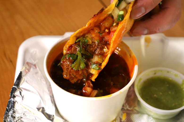 A birria taco being dipped in a cup on consomme from Taco de Birria by Rice Bar in